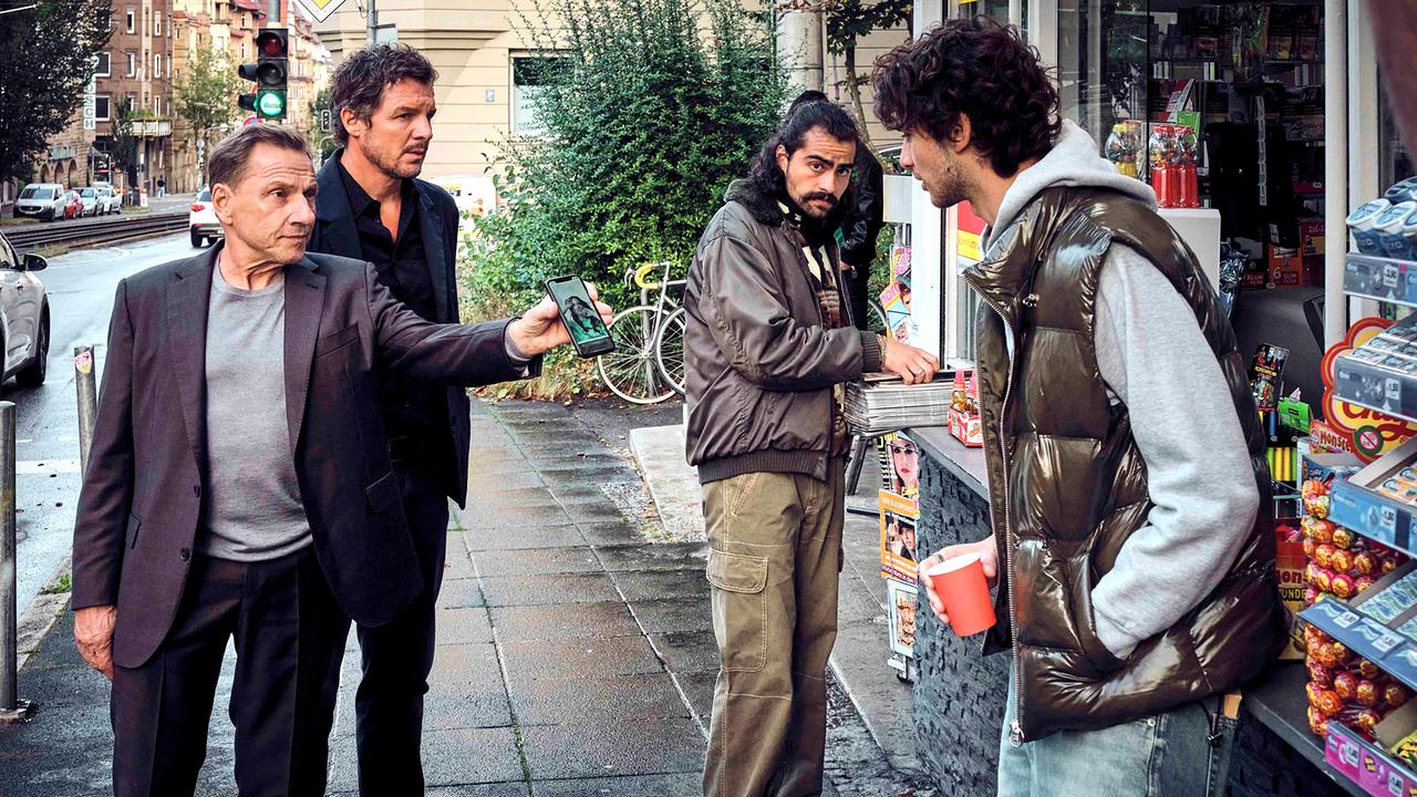 Thorsten Lannert und Sebastian Bootz zeigen zwei jungen Männern vor einem Kiosk ein Foto auf einem Smartphone.
