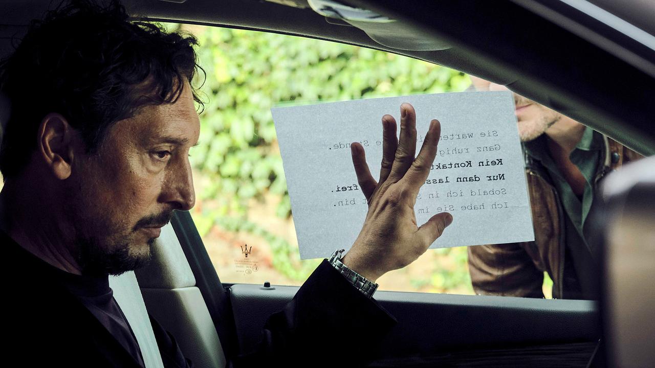Stephan Hübner im Auto hält einen Erpresserbrief an die seitliche Scheibe, den jemand von außen liest.
