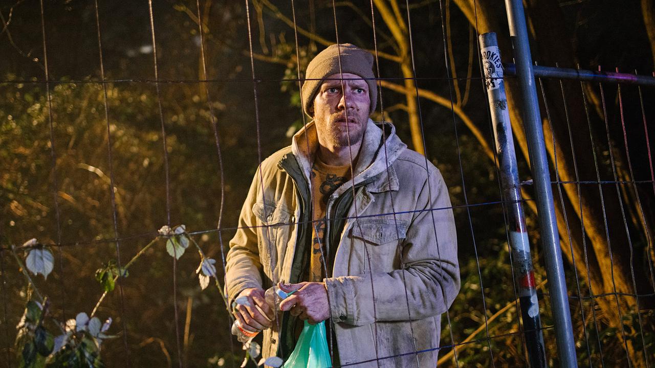 Der Obdachlose Emil Klaßen steht nachts hinter einem Bauzaun im Wald und hält eine Plastiktüte sowie eine Flasche.