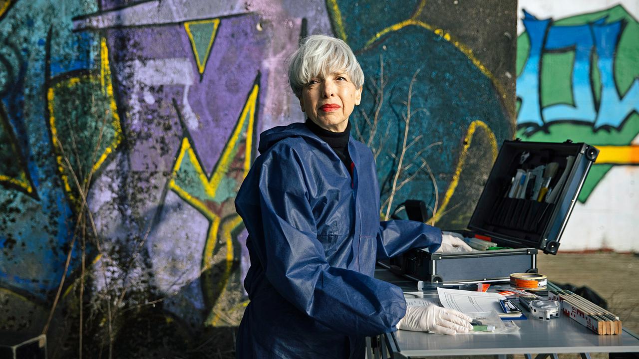 Gerichtsmedizinerin Edda Bingley in Schutzanzug und Handschuhen arbeitet mit forensischem Equipment vor einer bunt besprühten Wand.