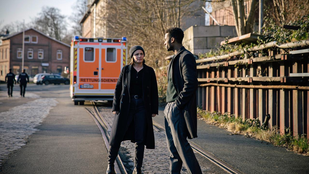 Liv Moormann spricht mit dem KDD Kollegen Patrice Schipper auf einer Straße vor einem Rettungswagen.