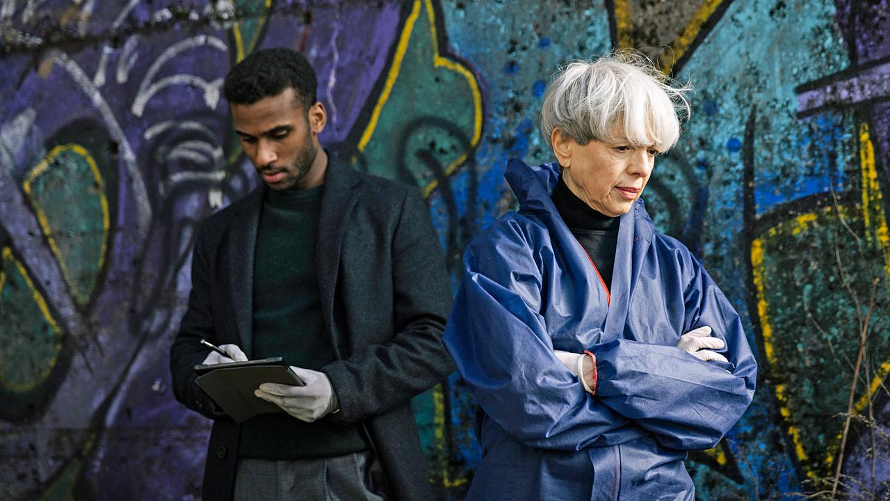 Patrice Schipper und Gerichtsmedizinerin Edda Bingley untersuchen den Tatort vor einer bunt besprühten Graffiti-Wand.