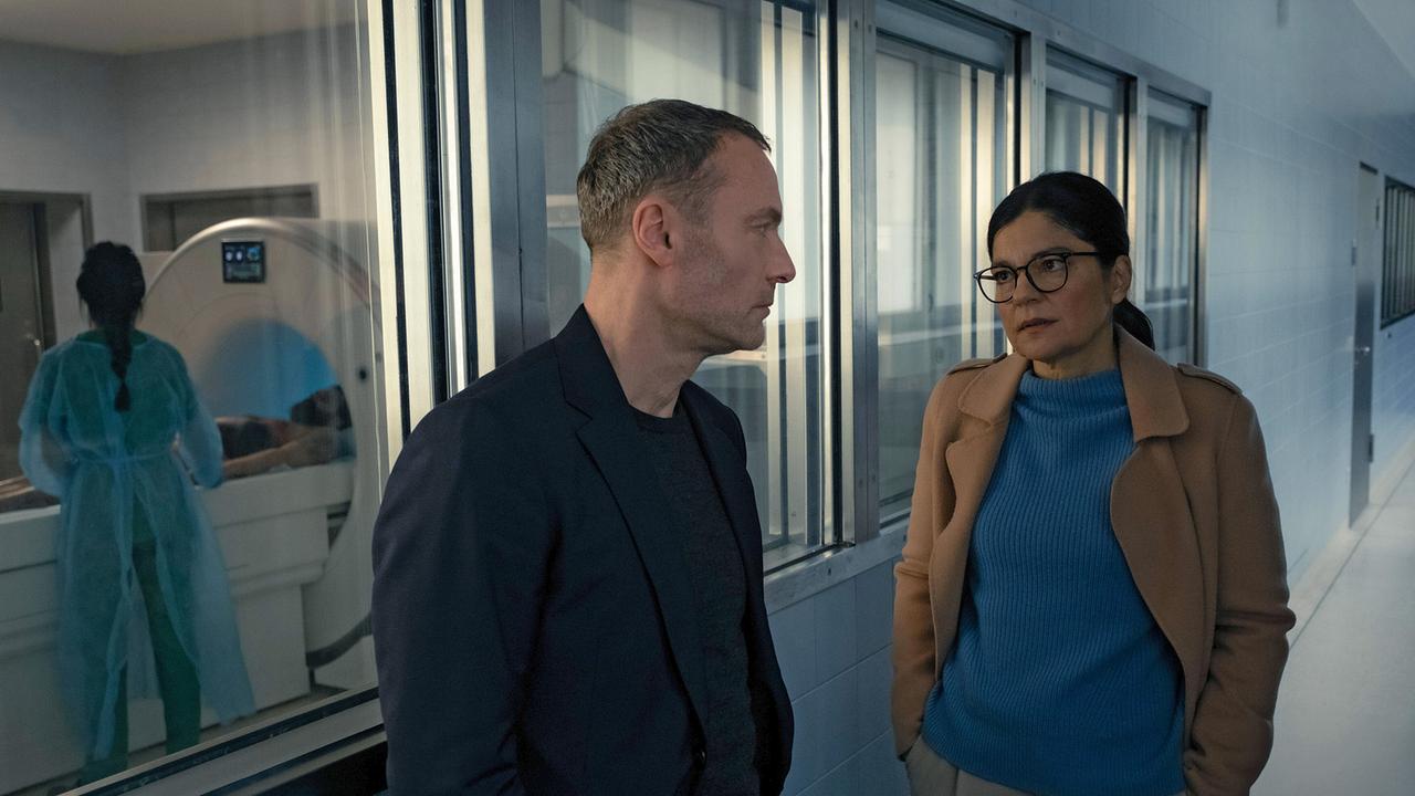 Staatsanwältin Sara Taghavi befragt Robert Karow zum Verbleib seiner verschollenen Kollegin, in einem Krankenhausflur vor einem Raum mit einem medizinischen Gerät, in dem jemand untersucht wird.