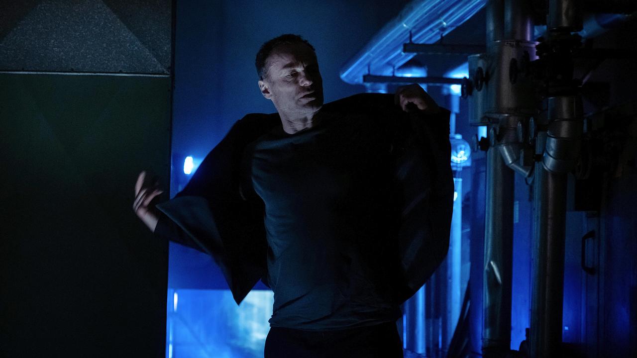 Robert Karow wirft sich in einem dunklen Raum mit blauem Licht eine Jacke über.