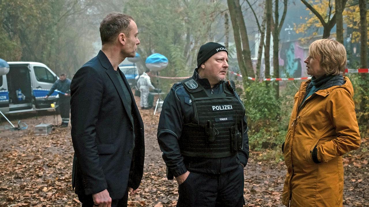 Susanne Bonard und Robert Karow stehen mit Polizeihauptmeister Salitter, am Tatort an einem abgesperrten Waldstück.