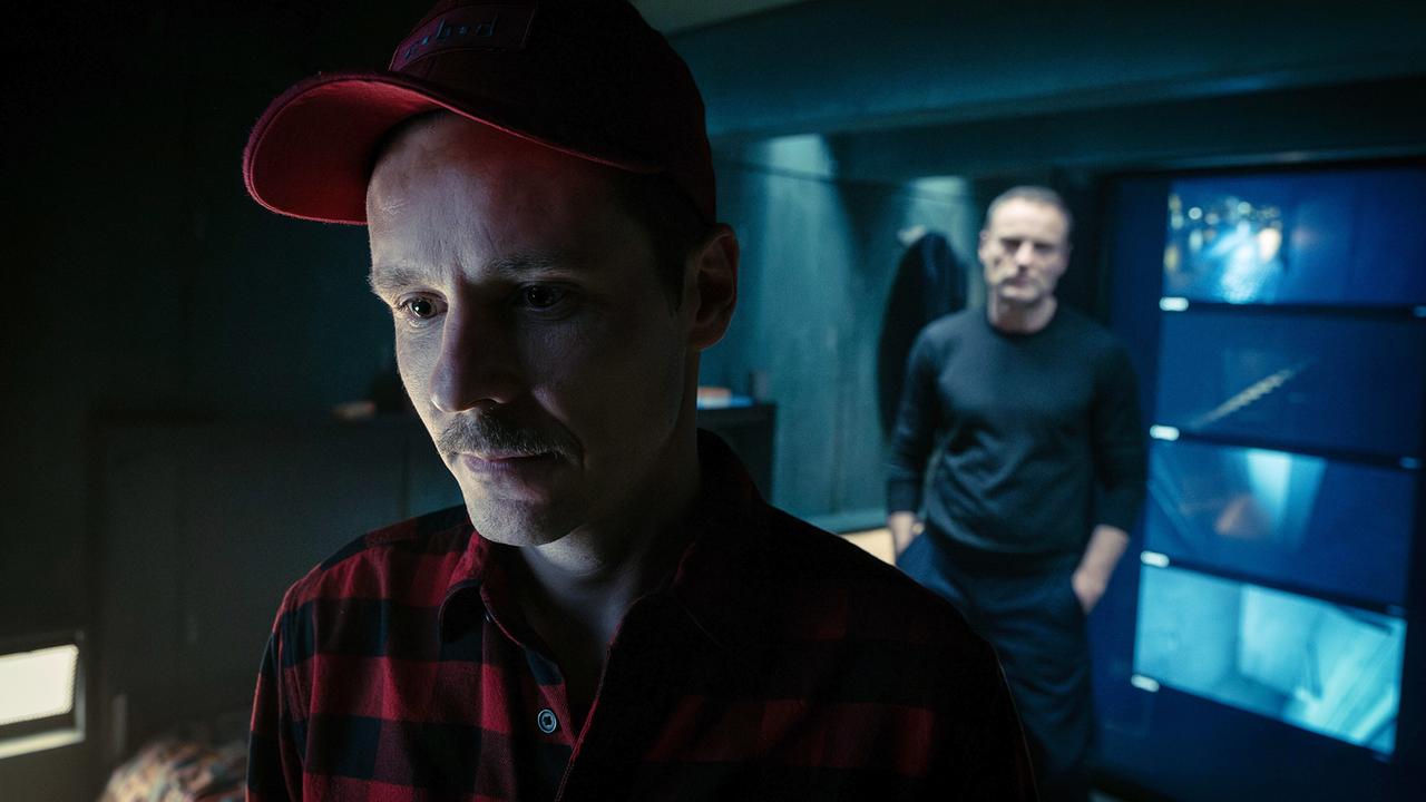 Robert Karow steht hinter dem Wildniswanderer Noah Farrell in einem dunklen Raum mit blaubeleuchteten Monitoren.