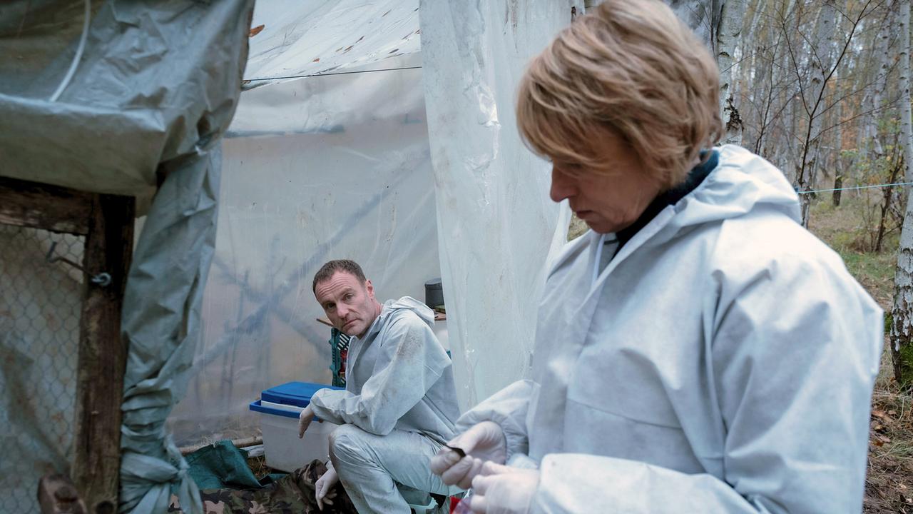 Susanne Bonard und Robert Karow, beide in Schutzanzügen, werten die Spuren am Tatort aus, wo der tote Obdachlose im Wald entdeckt wurde.