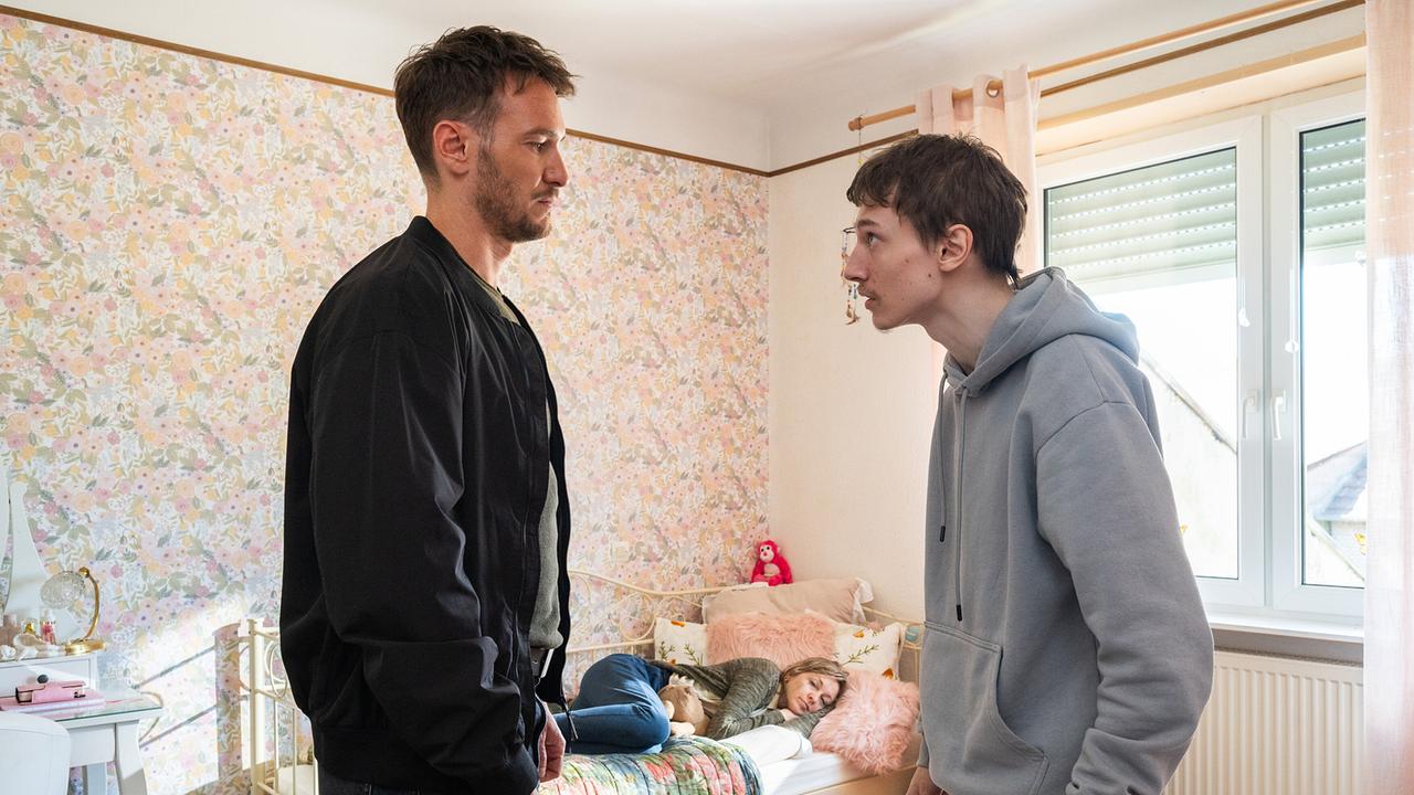 Leo Hölzer und Peter  Feidt stehen sich angespannt in einem Kinderzimmer gegenüber, während im Hintergrund Claudia Feidt auf dem Bett liegt.