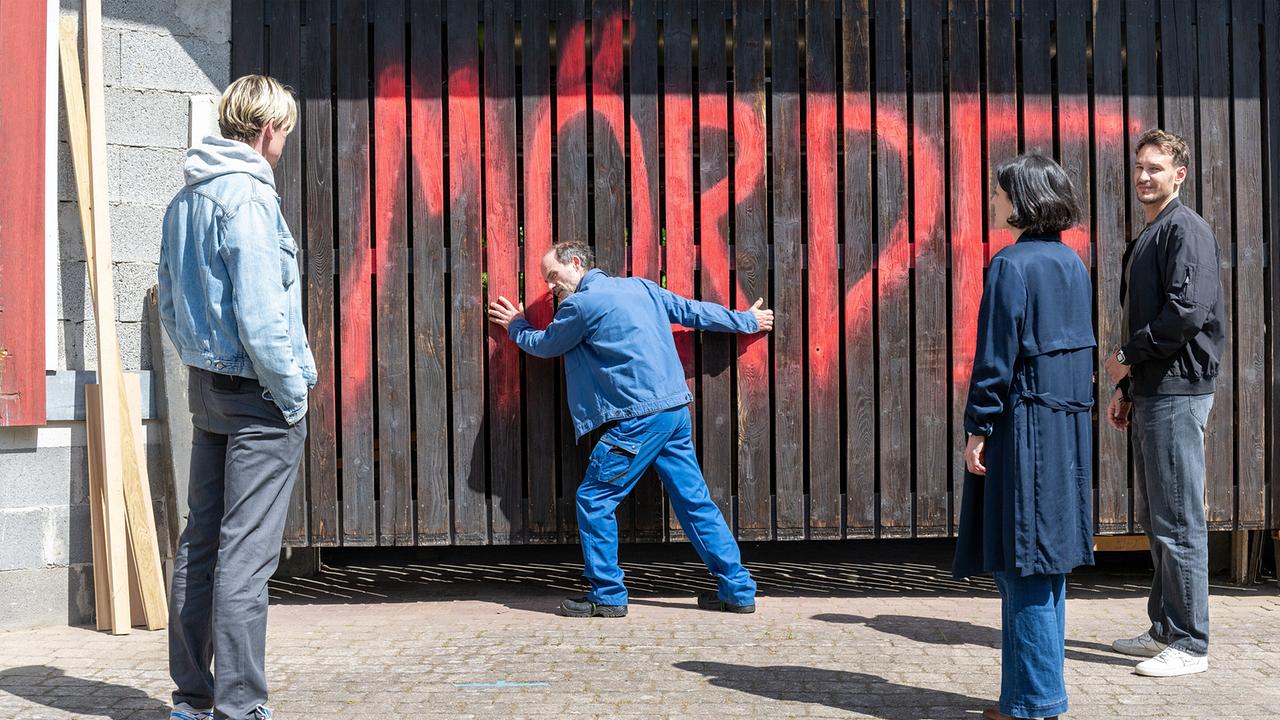 Adam Schürk, Esther Baumann, Leo Hölzer und Michel Louis stehen vor einem großen Holztor, auf dem in roter Farbe das Wort "Mörder" gesprüht wurde.