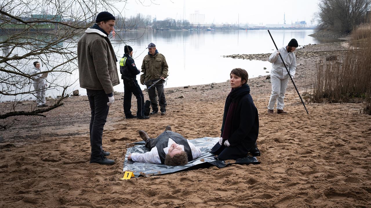Ermittlerinnen und Ermittler untersuchen am Ufer einen zugedeckten Körper und suchen nach Spuren. Elena Weber und ihr Kollege Jochen bemerken dabei, dass sie observiert werden.