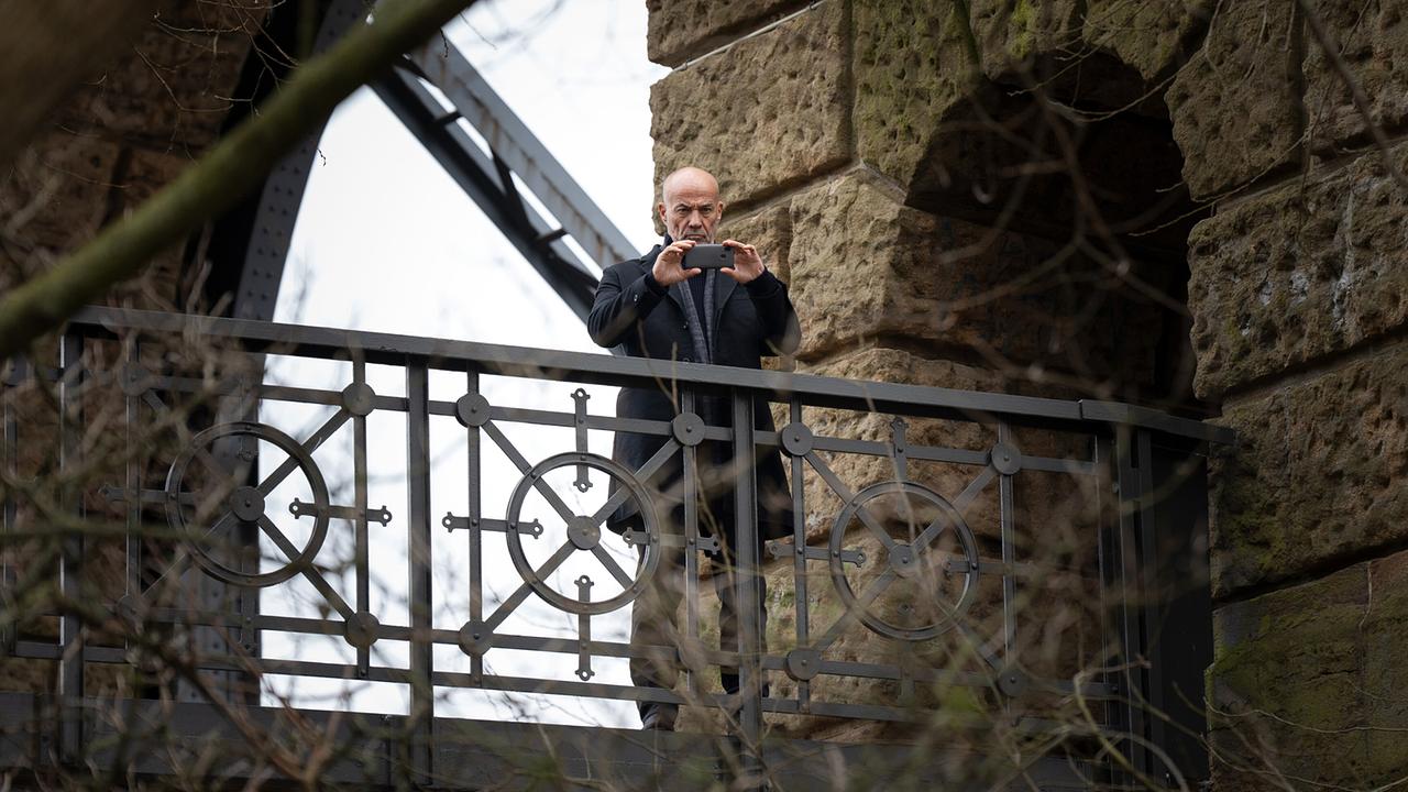Der ehemalige Kommissar Hagen Benz steht auf einer alten Brücke und fotografiert mit einem Smartphone fotografiert den Tatort aus der Ferne.