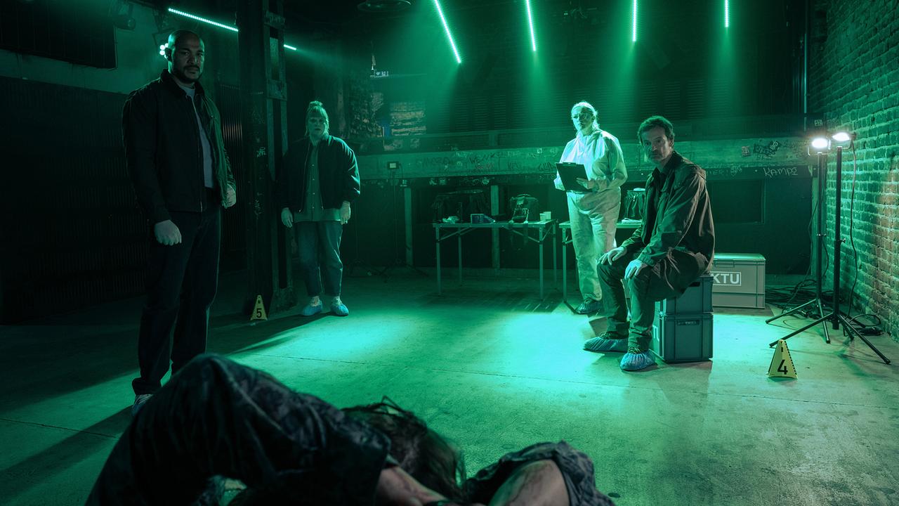 In einem grün ausgeleuchteten Raum begutachten Otto Pösken, Rosa Herzog, Greta Leitner  und Peter Faber die Leiche des Club-Managers Klaas Martens