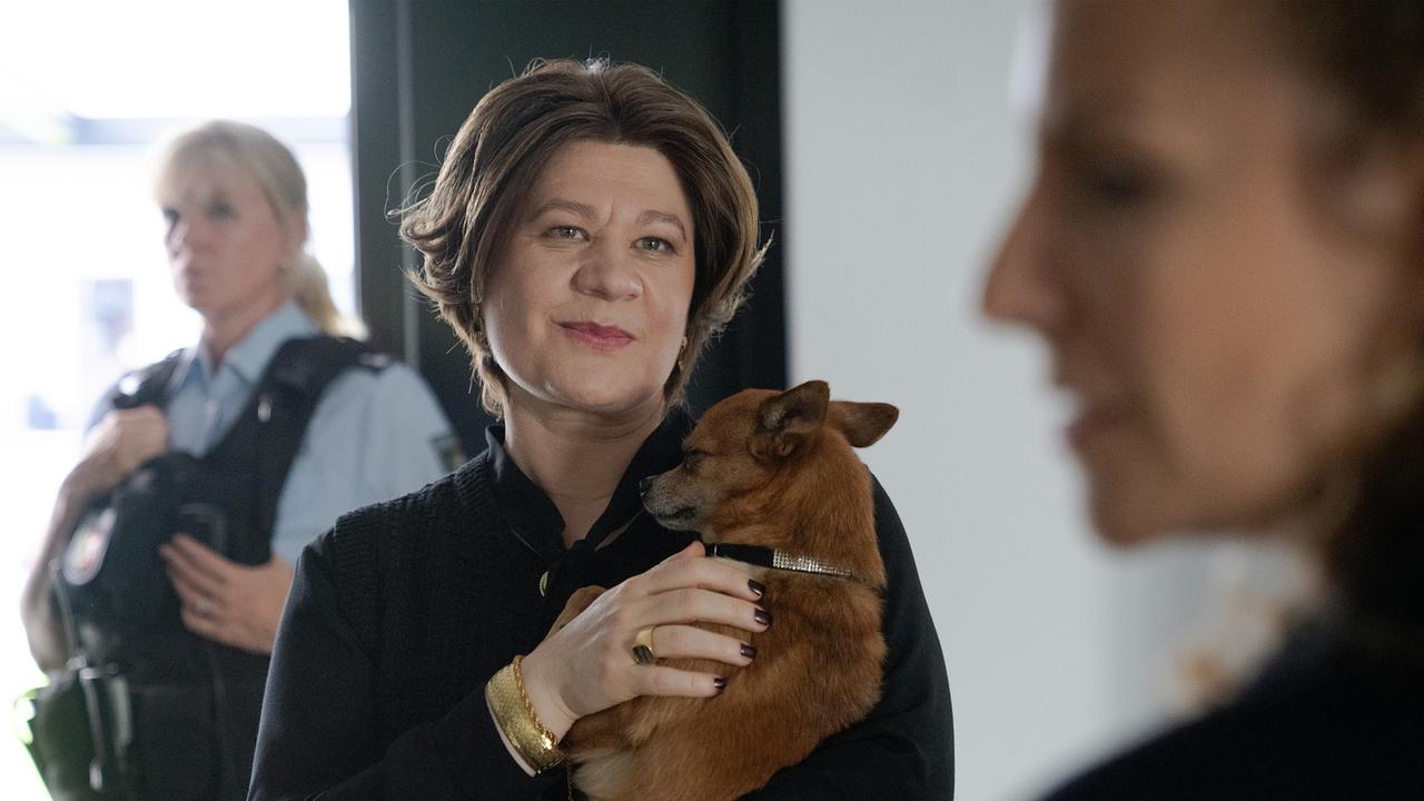 Klea Duka hält beim Gespräch mit Ira Klasnić in einem Innenraum einen kleinen Hund im Arm, während im Hintergrund eine Polizistin steht.