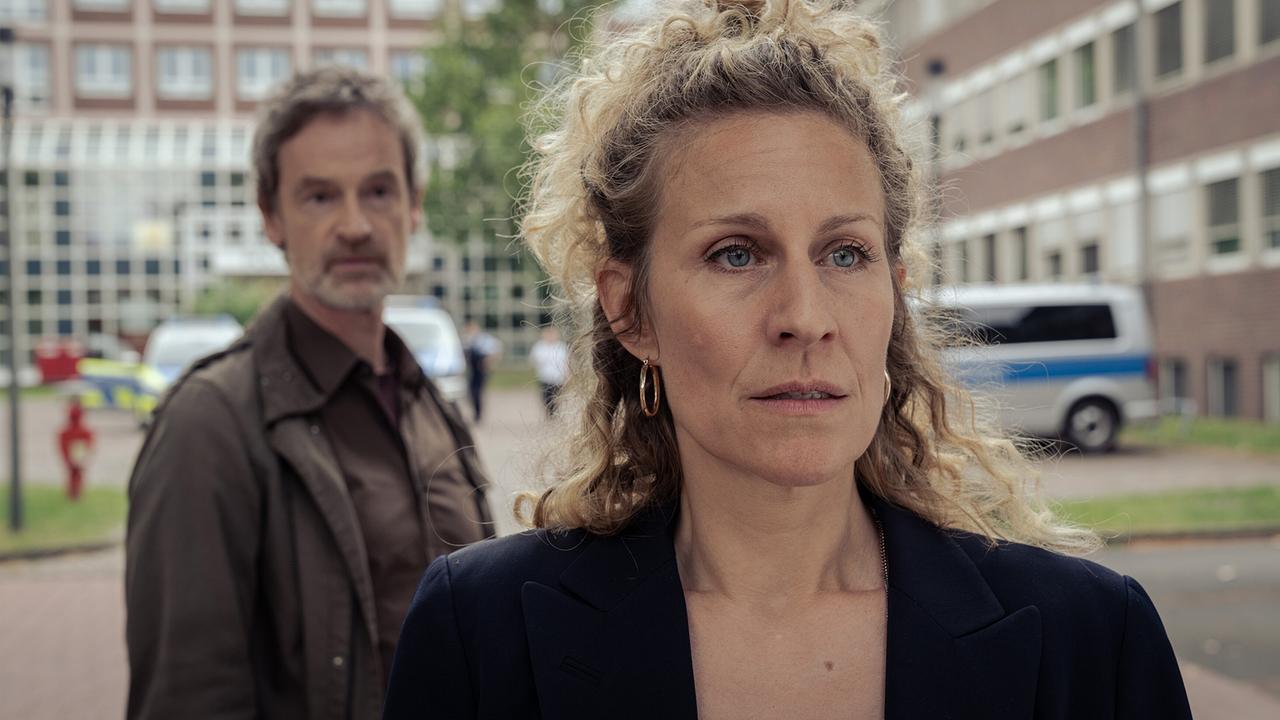 Ira Klasnić steht vor Peter Faber draußen vor einem Gebäude, im Hintergrund ist ein Polizeifahrzeug zu sehen.