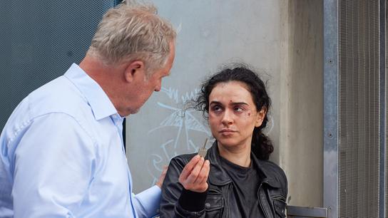 Die im Gesicht verletzte Azra zeigt Moritz Eisner einen Schlüssel im Freien vor einer Wand.