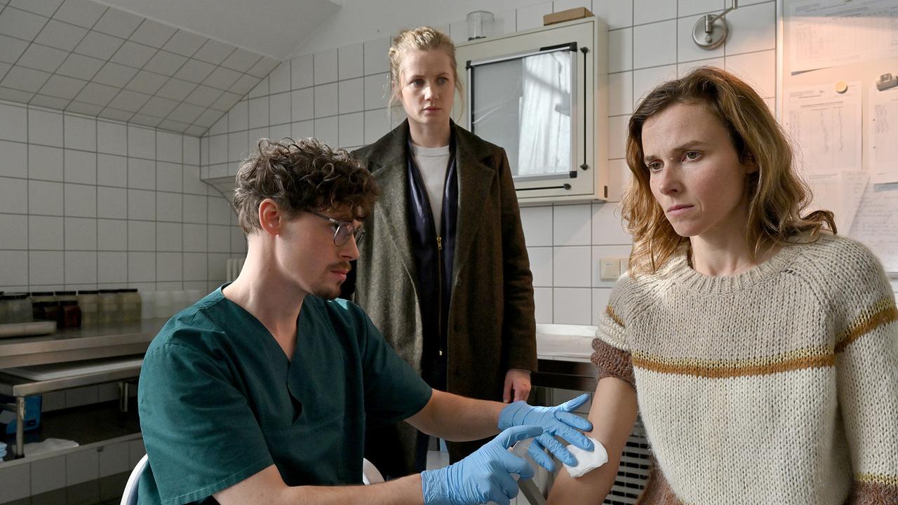 "Tatort: Unsichtbar": Karin Gorniak (Karin Hanczweski) lässt sich von Gerichtsmediziner Jonathan Himpe (Ron Helbig) Blut abnehmen, um die Probe mit dem Blut des Opfers vergleichen zu können, im Hintergrund Leo Winkler (Cornelia Gröschel).