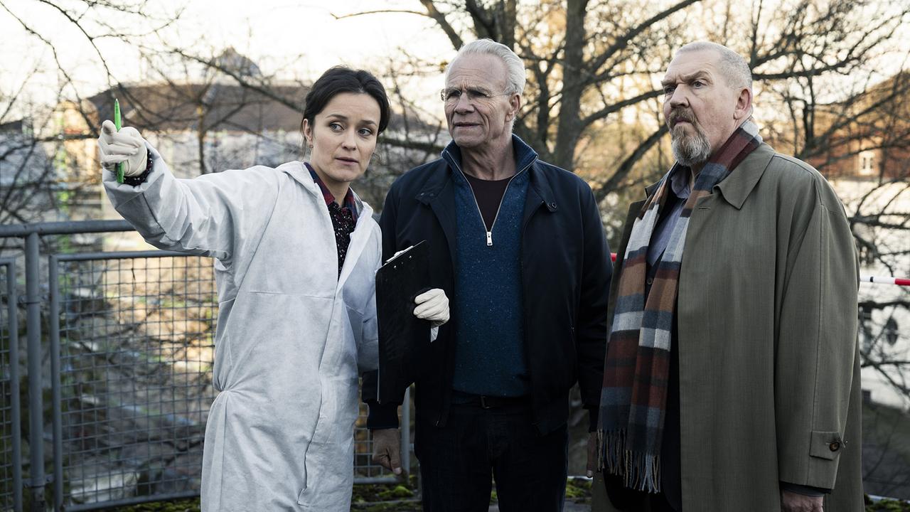 Im Freien erklärt KTU-Chefin Natalie Förster in Schutzkleidung Max Ballauf und Freddy Schenk etwas und zeigt dabei in die Umgebung.