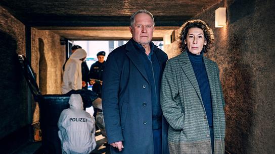 Moritz Eisner und Bibi Fellner stehen in einem Durchgang, im Hintergrund arbeiten Polizeibeamte und die Spurensicherung.