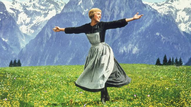 Julie Andrews dreht sich als Maria in schwarzem Kleid und gestreifter Schürze mit ausgebreiteten Armen auf einer blühenden Almwiese vor einer Kulisse aus hohen, schneebedeckten Bergen