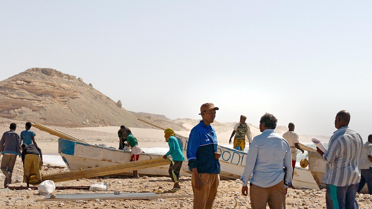 Mehrere Männer, unter ihnen Mamargade, arbeiten rund um einfache Holzboote, die auf den Strand gezogen wurden in einer sandigen Landschaft mit Meer im Hintergrund.