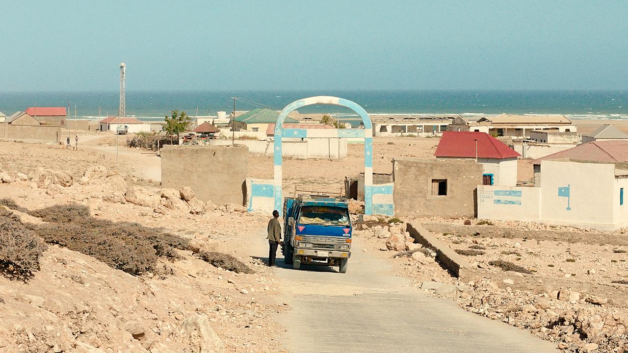 Ein kleines Areal an der Küste mit einfachen Häusern liegt an einer staubigen Straße nahe dem Meer. Vor einem Torbogen steht ein Lieferwagen mit einem Mann daneben.