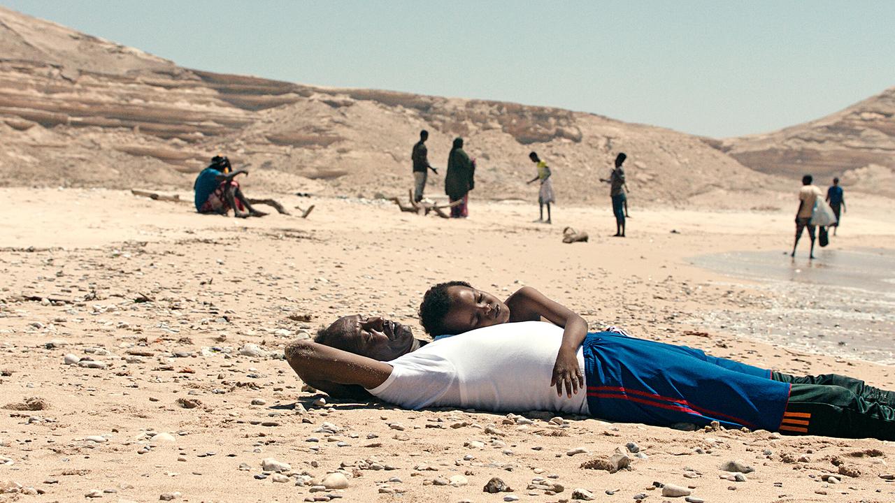 Mamargade liegt mit Cigaal, der sich an ihn kuschelt, am Strand, während andere Menschen im Hintergrund stehen.