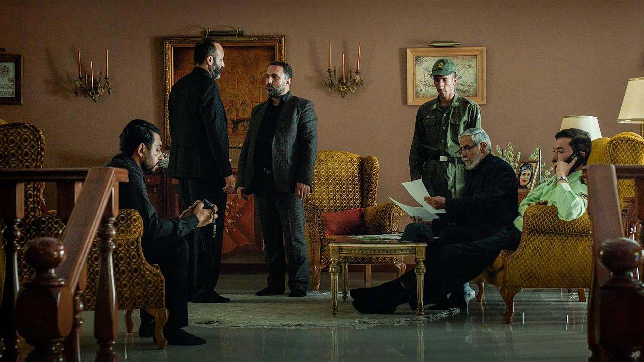 Mehrere Männer, darunter ein Soldat und ein Sicherheitsoffizier, besprechen sich in einem opulent eingerichteten Wohnzimmer, während einer telefoniert.