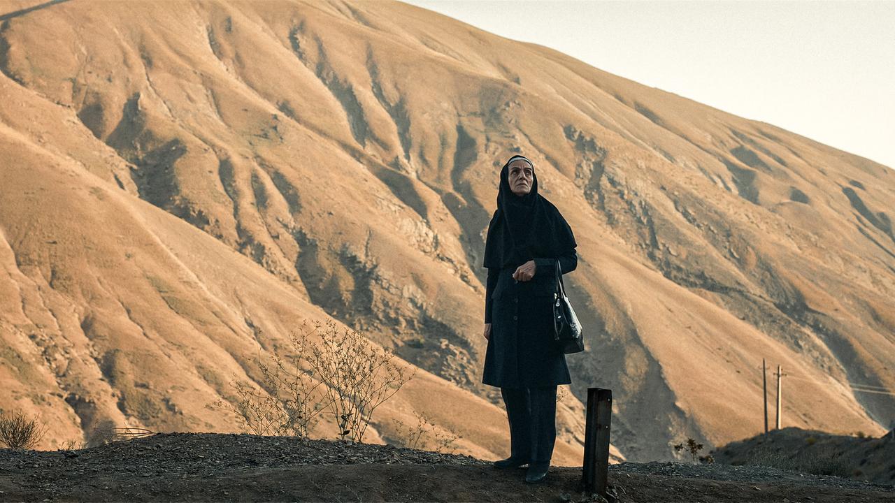 Tarlan mit schwarzem Kopftuch steht allein vor einer kargen, bergigen Landschaft.