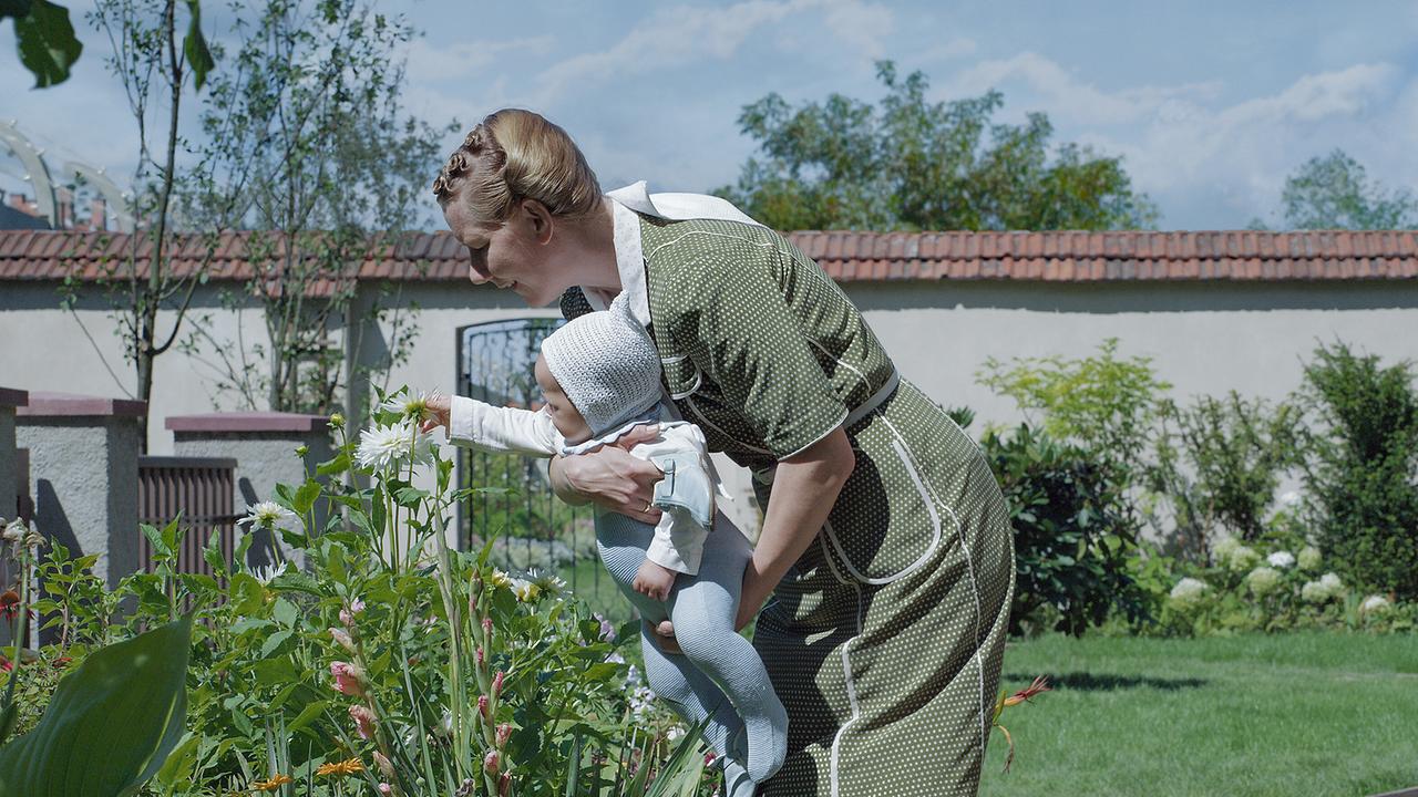 In einem Garten, der von einer Mauer abgegrenzt ist, steht Sandra Hüller als Hedwig Höß, die ihr Baby hält, damit es eine Blume anfassen kann