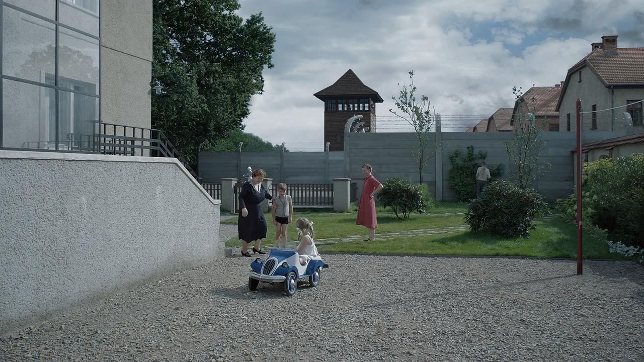 Im Garten der Familie Höß, Hedwig mit ihrer Mutter mit den Kindern, die mit einem Tretauto spielen, hinter der unverputzten Mauer ist ein Wachturm und große Gebäude zu sehen