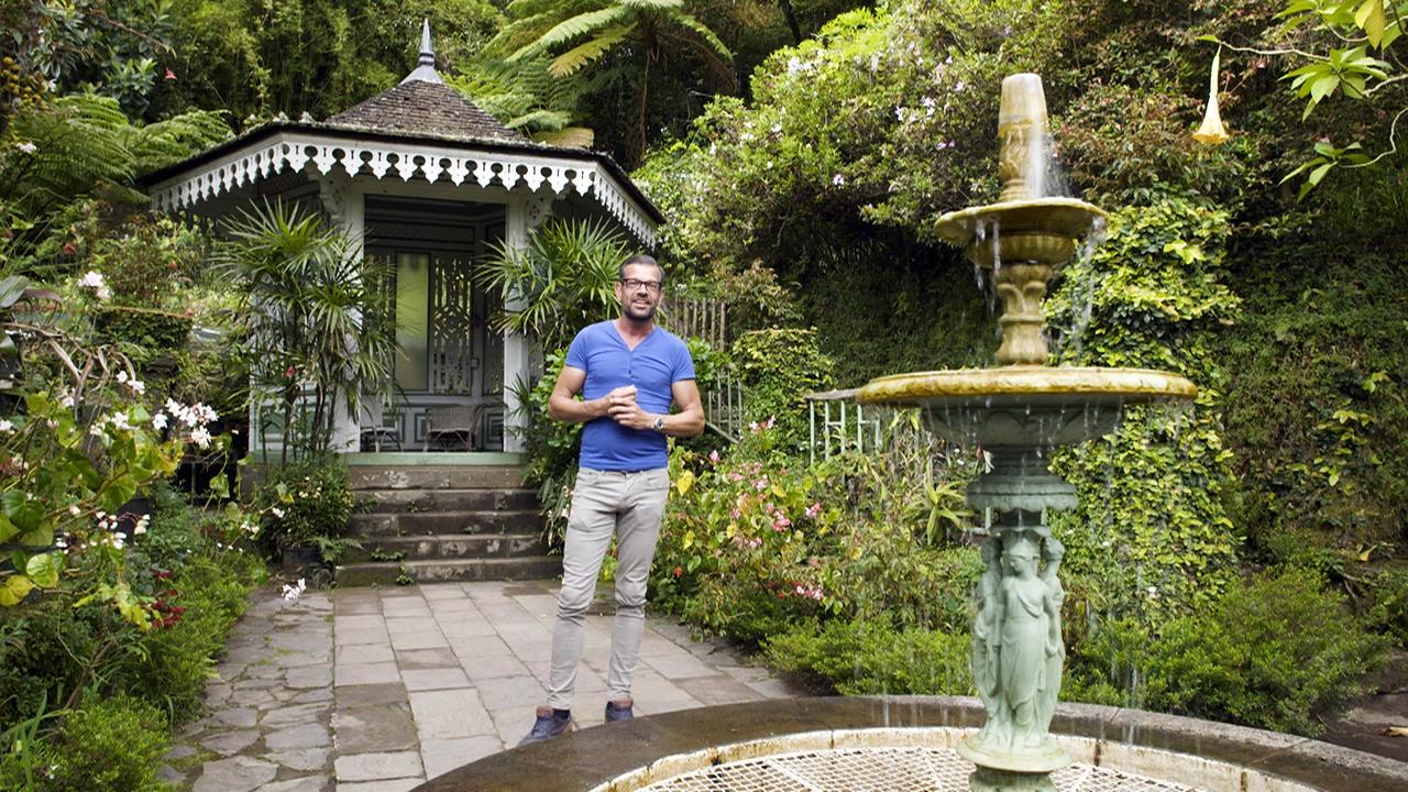 Karl Ploberger steht in einem Garten vor einem steinernen Springbrunnen mit Figuren und einem kleinen Pavillon mit Spitzdach im Hintergrund