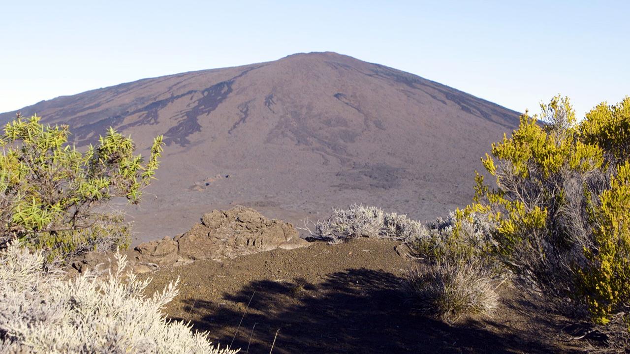 Der aktive Vulkan „Piton de la Fournaise“ umgeben von grünen und grauen Sträuchern im Vordergrund