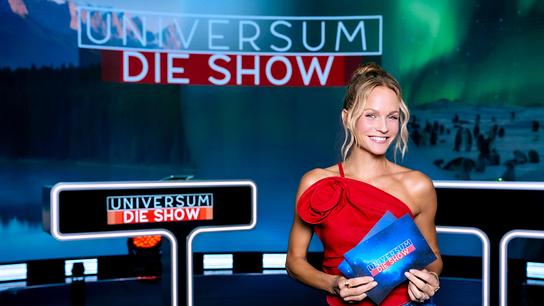 Mirjam Weichselbraun in rotem Top präsentiert sich im „Universum – Die Show“-Studio und hält Karten der Sendung.