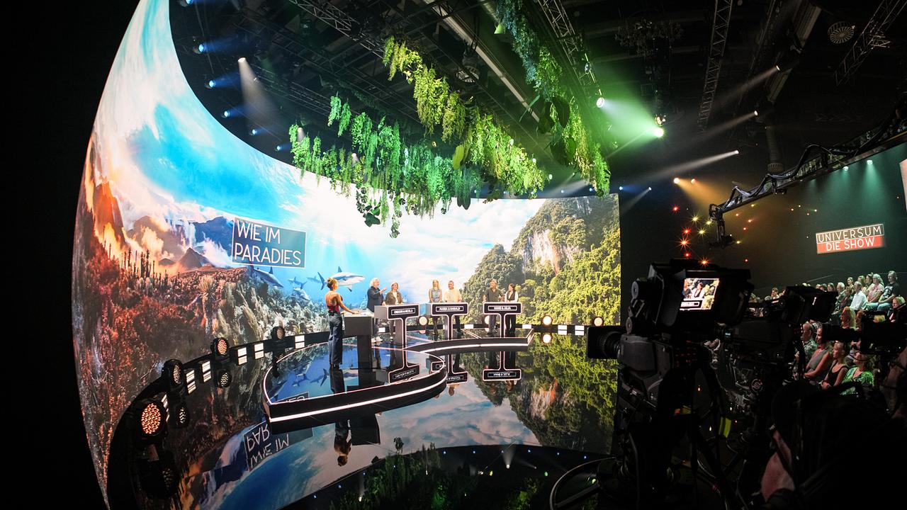 Mirjam Weichselbraun, Gregor Seberg, Nicola Feichtegger, Maria Angelini-Santner, Harald Zeitlhofer, Hans Knaus und Julia Lauss stehen an ihren Spielpulten im farbenfrohen Studio von „Universum – Die Show“.