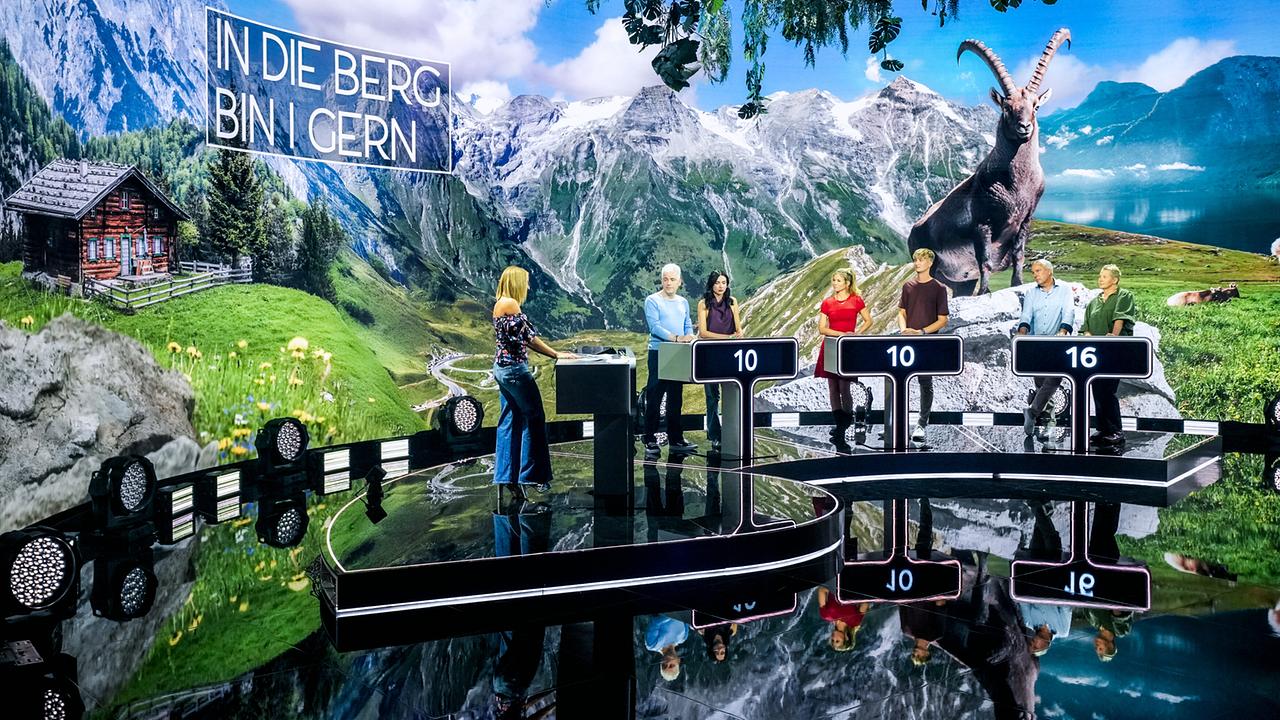 © ORF/Roman Zach-Kiesling Mirjam Weichselbraun steht mit Michael Mittermeier, Anna Reichenpfader, Hilde Dalik, Marius Schneider, Fritz Strobl, Brigitte Treichl-Krätchmer im "Universum - Die Show"-Studio vor einer imposanten Alpenlandschaft mit Steinbock und dem Schriftzug „In die Berg bin i gern“.