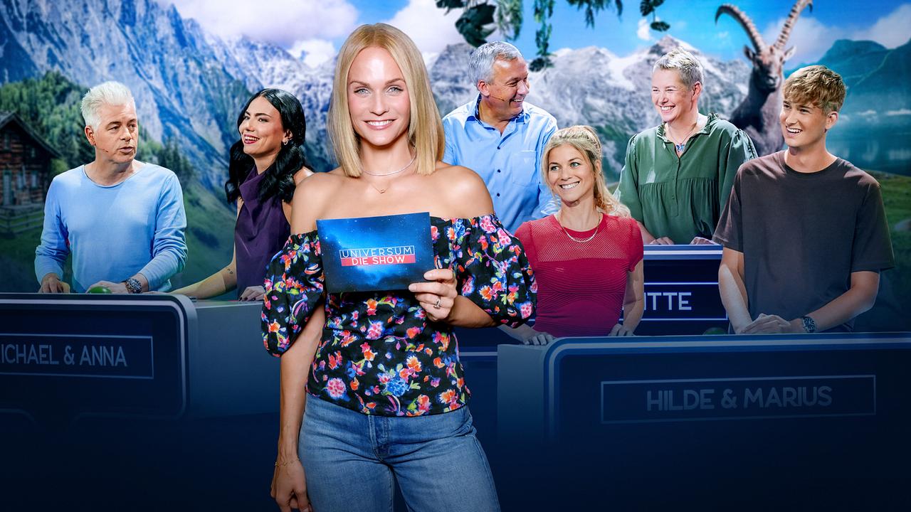 Mirjam Weichselbraun steht lächelnd im Fernsehstudio zu „Universum – Die Show“, im Hintergrund sind Michael Mittermeier, Anna Reichenpfader, Fritz Strobl, Brigitte Treichl-Krätschmer, Hilde Dalik und Marius Schneider an den Spielpulten zu sehen.
