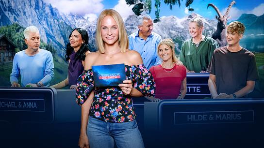 © ORF/Roman Zach-Kiesling Mirjam Weichselbraun steht lächelnd im Fernsehstudio zu „Universum – Die Show“, im Hintergrund sind Michael Mittermeier, Anna Reichenpfader, Fritz Strobl, Brigitte Treichl-Krätschmer, Hilde Dalik und Marius Schneider an den Spielpulten zu sehen.