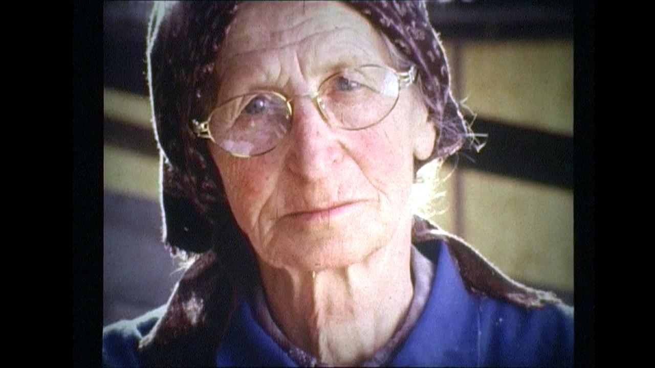 Die ältere Elisabeth Sitter mit Kopftuch und Metallbrille ist in einer körnigen, farbstichigen Filmaufnahme im Porträtstil zu sehen.