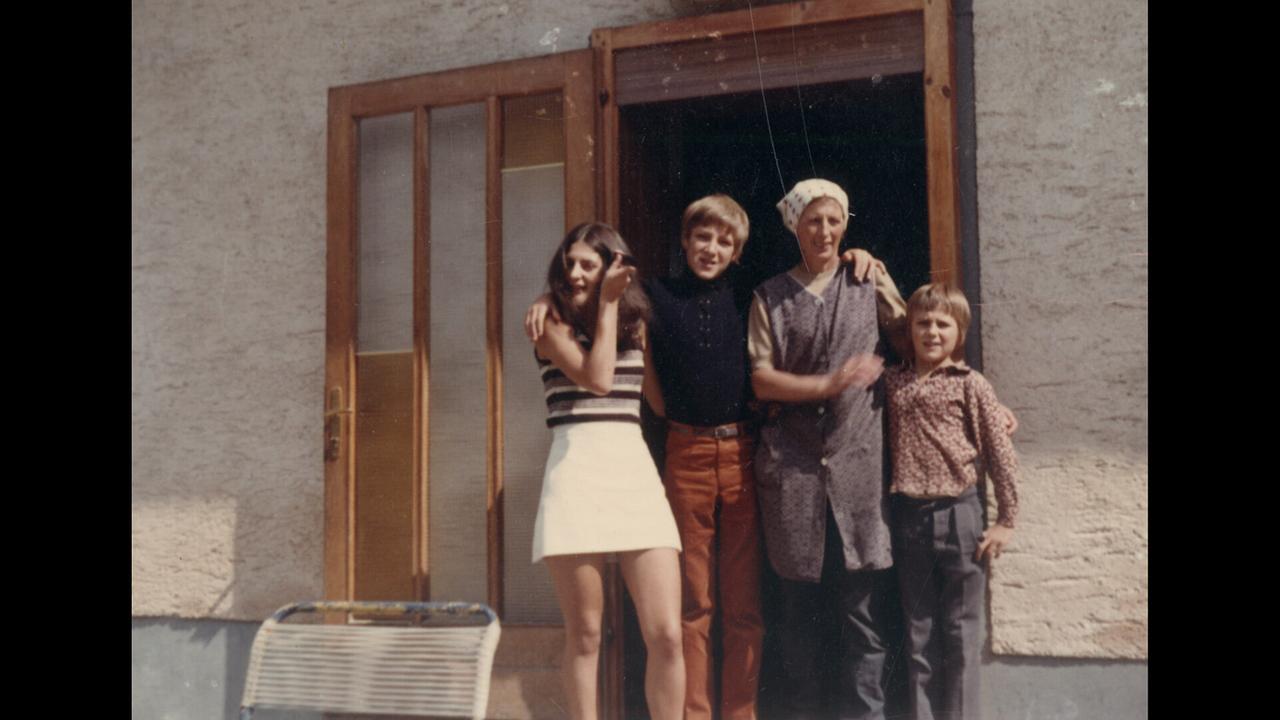 Die Familie Mračnikar in den 70er Jahren: Vier Menschen stehen Arm in Arm vor einer Haus­tür und posieren für ein Familienfoto.