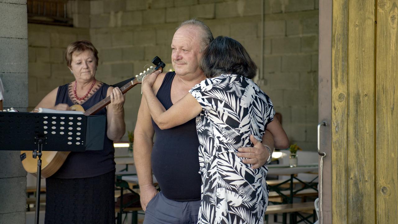 Eugen und Lizi Sitter eng umschlungen in einem offenen, rustikal wirkenden Raum, während daneben eine Musikerin auf einer Gitarre spielt.