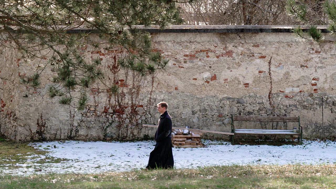 © ORF Frater Johannes Neuß steht im Schnee vor einer an einer alten Steinmauer, an der eine einfache Bank im Hintergrund zu sehen ist.