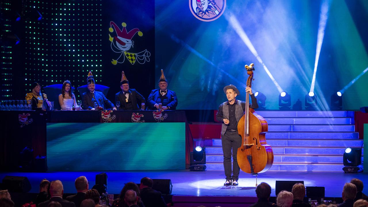© ORF/Peter Krivograd "Die Spaßbrems`n" Arnold Angermann steht mit Kontrabass auf der blau ausgeleuchteten Bühne, während im Hintergrund das Villach‑Wappen und eine Sitzreihe zu sehen sind.