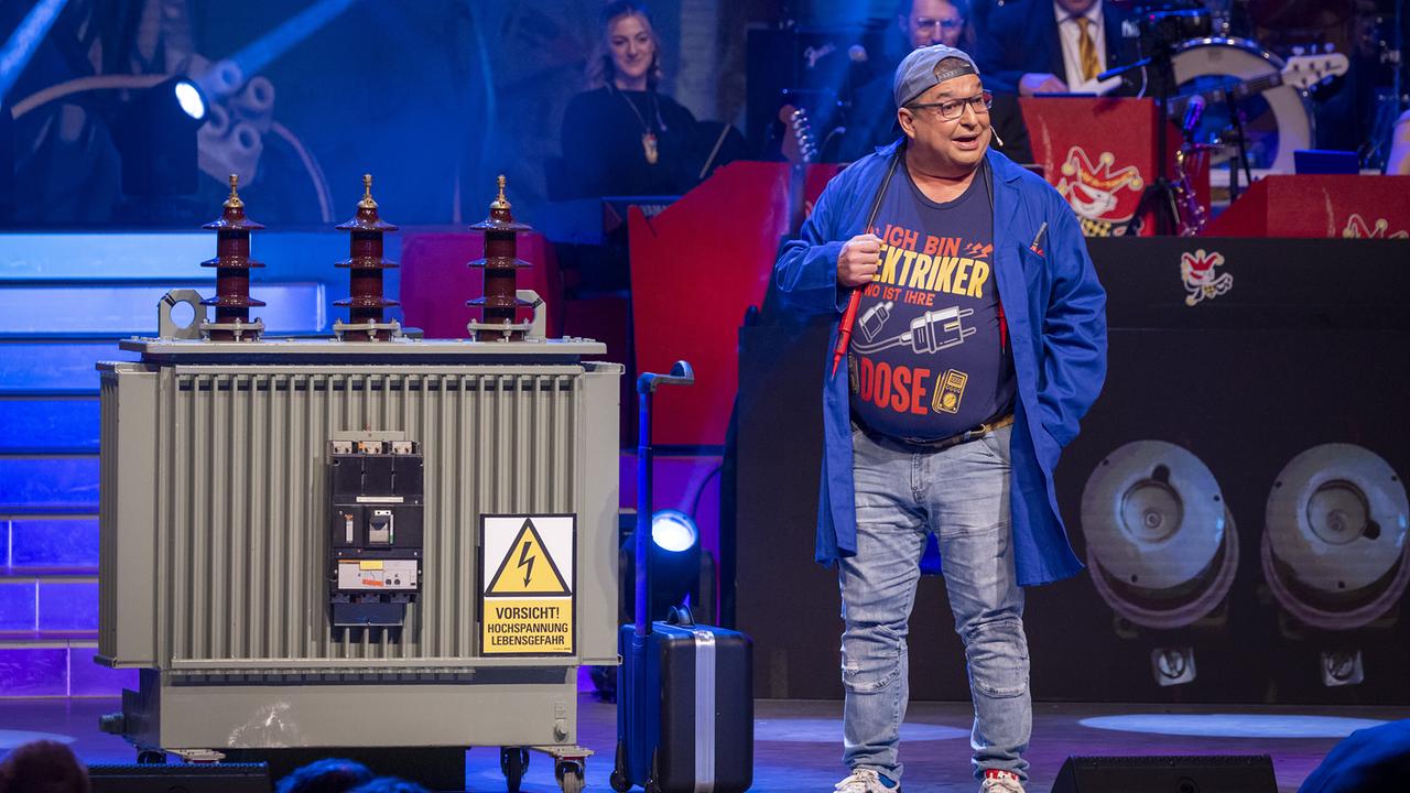 © ORF/Peter Krivograd Herbert Rader als Der Elektriker steht im Arbeitskittel mit Mikrofon neben einem großen Transformator mit Warnschild „Hochspannung“ auf einer Showbühne.