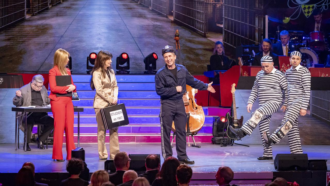 © ORF/Peter Krivograd Mehrere Darsteller:innen darunter Arnold Angermann in Polizeiuniform spielen eine Bühnenszene, rechts marschieren zwei Männer in gestreiften Sträflingskostümen, links stehen zwei Frauen im Anzug.
