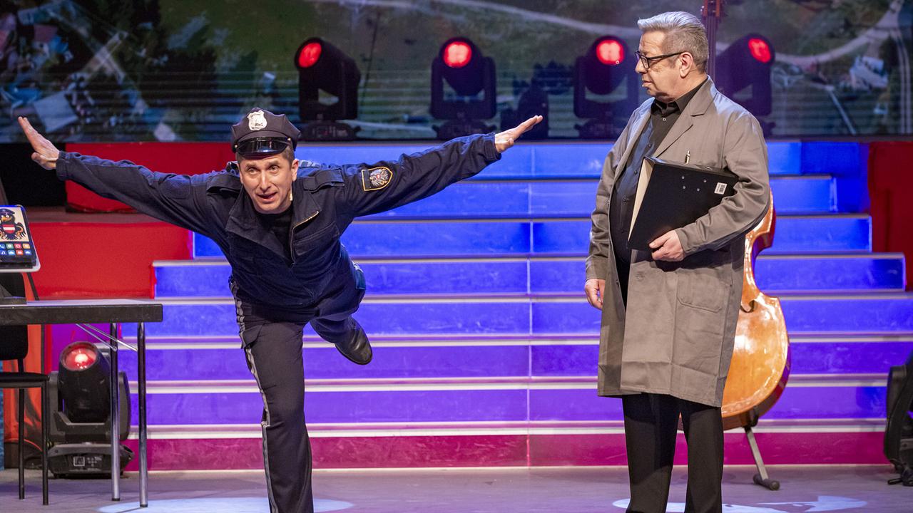 © ORF/Peter Krivograd Arnold Angermann in Polizeiuniform balanciert einbeinig mit ausgebreiteten Armen neben Hannes Krenner im grauen Mantel auf der Bühne.