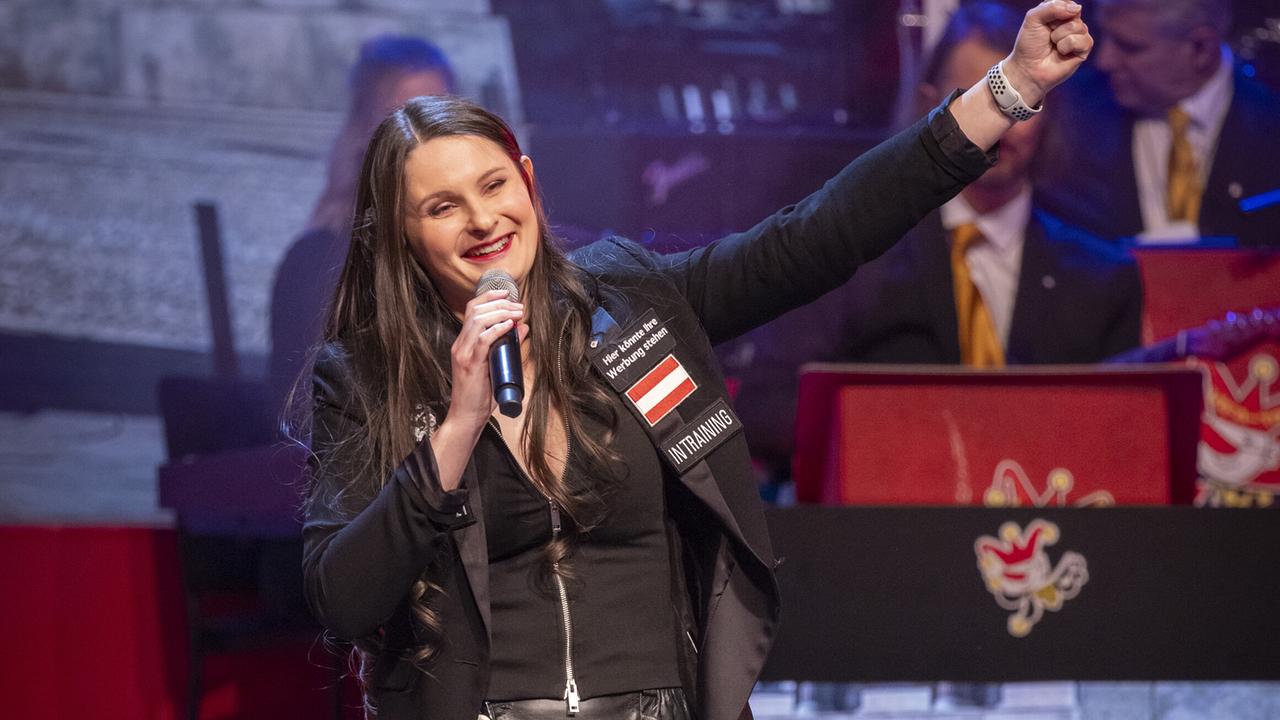 © ORF/Peter Krivograd Die Praktikantin Eva Mion spricht auf der Bühne ins Mikrofon und hebt die Hand, im Hintergrund sitzt eine Band.