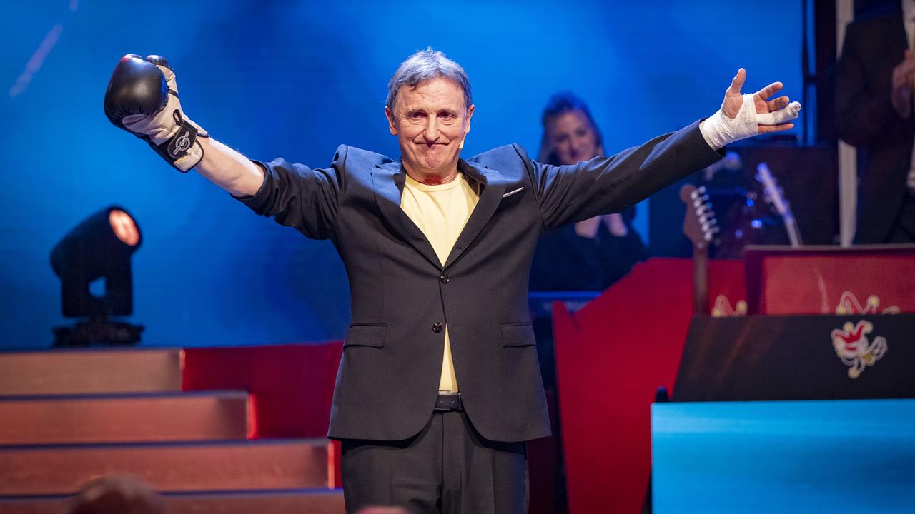 © ORF/Peter Krivograd "Noste" Manfred Obernosterer im Anzug steht mit Boxhandschuh und bandagierter Hand im Rampenlicht und breitet die Arme aus.