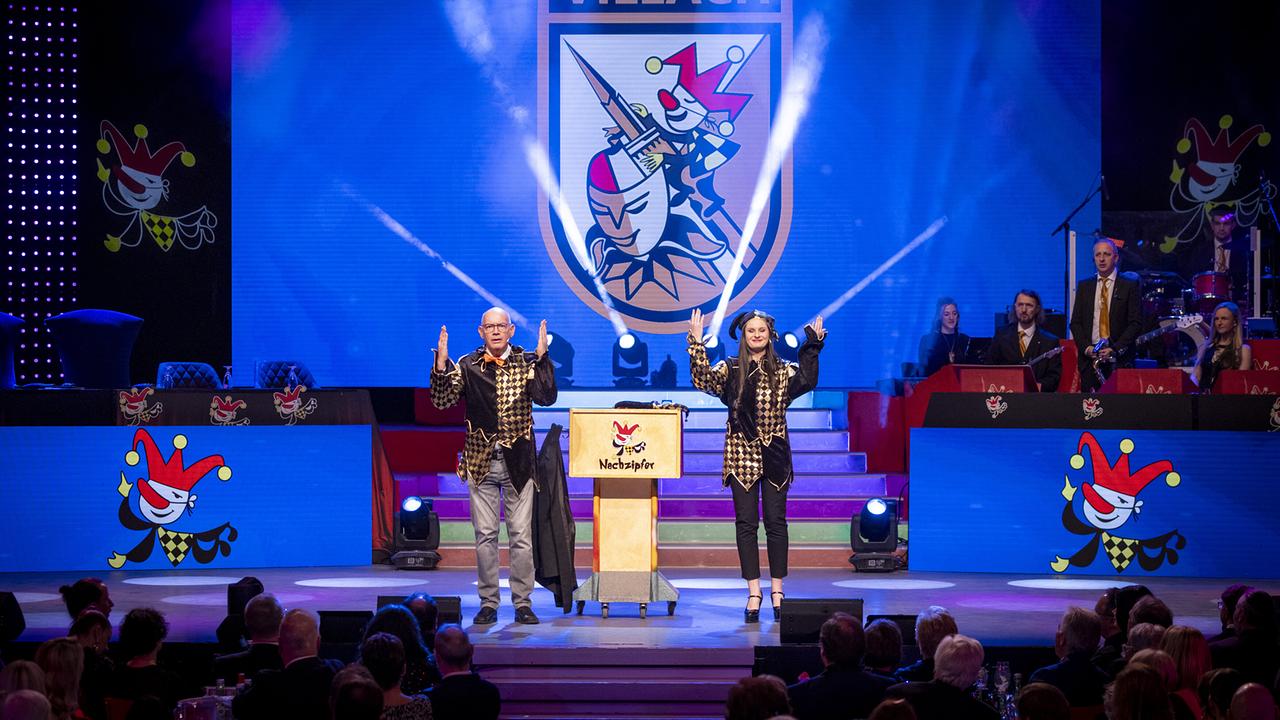 © ORF/Peter Krivograd "Narrzipfer" Hannes Höbinger und Eva Mion in harlekinartigen Kostümen stehen am Rednerpult vor dem großen Villach‑Wappen auf der Bühne.