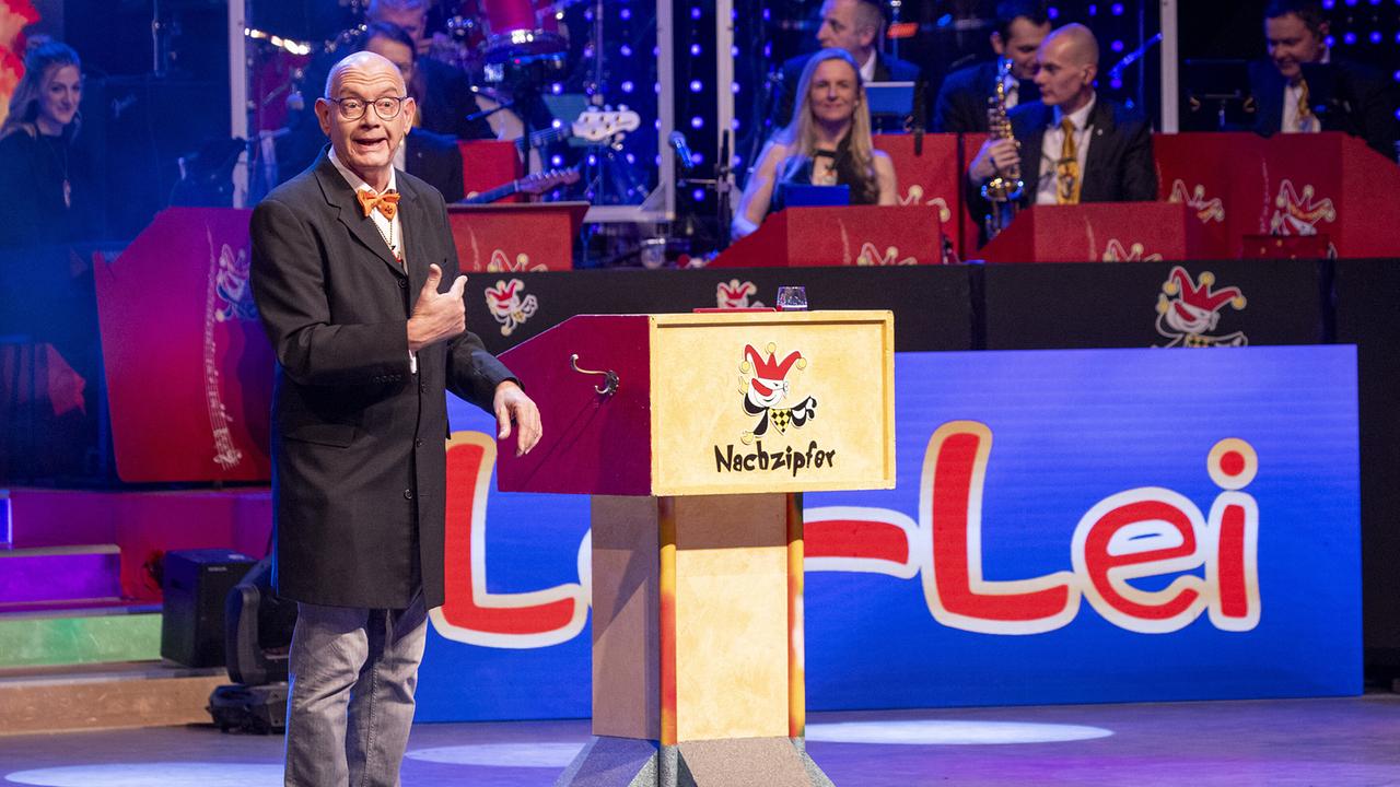 © ORF/Peter Krivograd "Narrzipfer" Hannes Höbinger im dunklen Mantel steht neben einem Rednerpult mit „Nachzipfer“-Logo, im Hintergrund sitzt die Band.