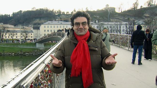© ORF/ORF-Salzburg Startenor und Mozartwochen-Intendant Rolando Villazon mit rotem Schal und Anorak steht auf einer Fußgängerbrücke, an deren Geländern zahlreiche Liebesschlösser hängen.